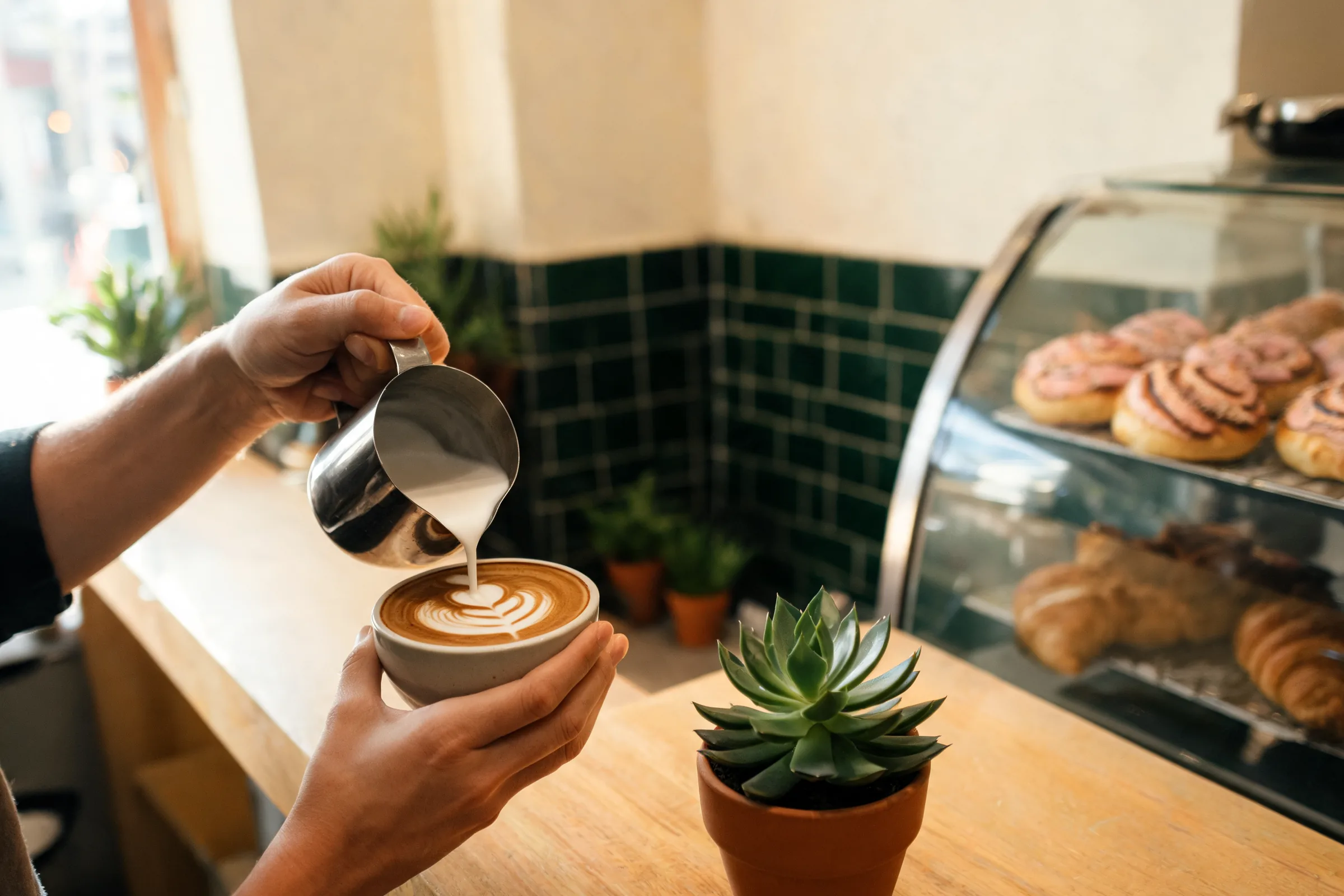Barista sirviendo un latte en la barra de una cafetería, con la vitrina de pan dulce al lado